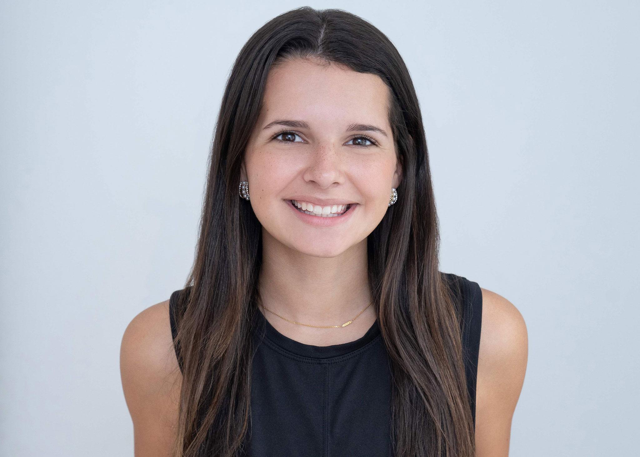 Kaitlin-Lawrence-Abbey-Brown-Novo-Fitness-Youth-Services Headshot of Kaitlin Lawrence, a Youth Services team member at Novo Fitness Studio. Kaitline wears a black tank top. Her long brown hair cascades over her shoulders as she smiles at the camera.