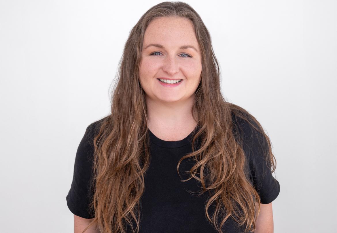Headshot of Christine, a smiling member of the Client Care Team at Novo Fitness Studio, wearing a black shirt with long wavy hair, in front of a white background.