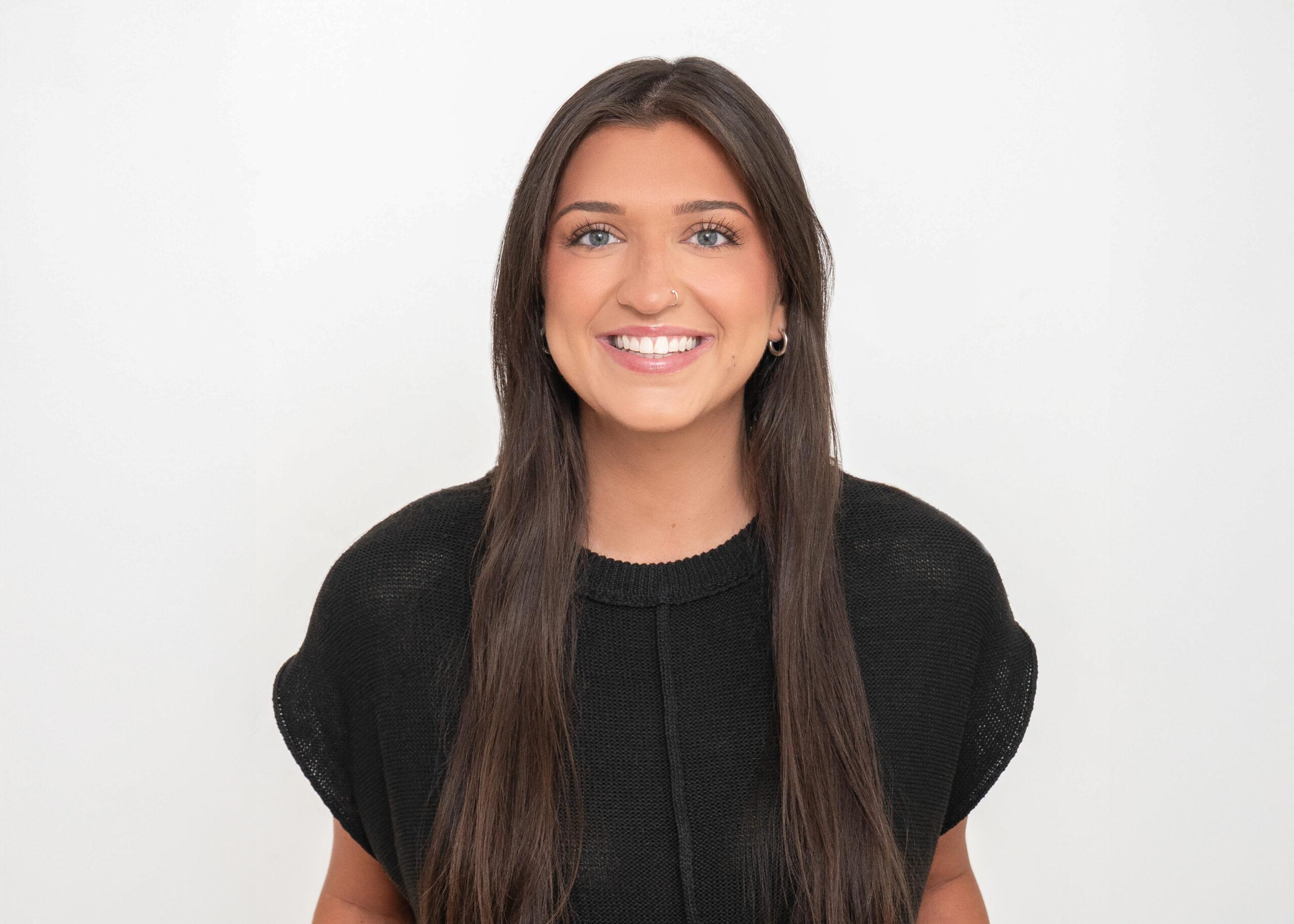 Headshot of Danica Rains wearing a black knit top, smiling with long straight brown hair, in front of a white background at Novo Fitness Studio in Columbus.